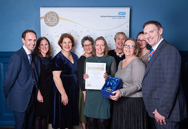 Nine smartly dressed smiling people, 7 women with a man at each side, holding a certificate and presentation box