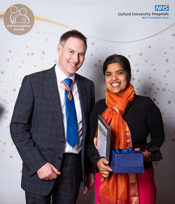 Smart man in suit and tie stands alongside lady in bright saree holding framed certificate and presentation box