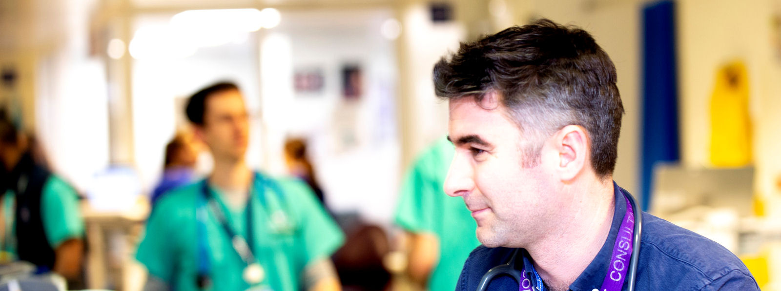 Man in scrubs, seated, busy clinical area behind