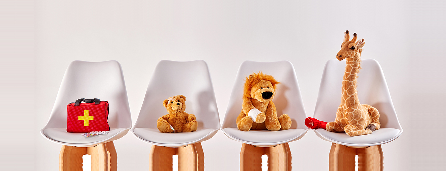 Four wiating room chairs with a toy medical bag, cuddly teddy, cuddly lion and cuddly giraffe placed on them, one on each