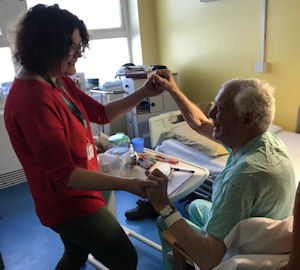 An elderly patient in a chair by a hospital bed is encouraged to dance by a young woman, holding his hands