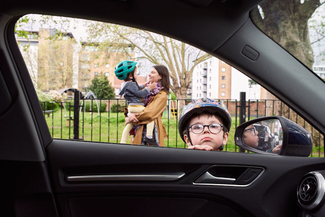 Child in cycle helmet peers through car window: behind is an urban playpark where a woman lifts another small child in a cycle helmet