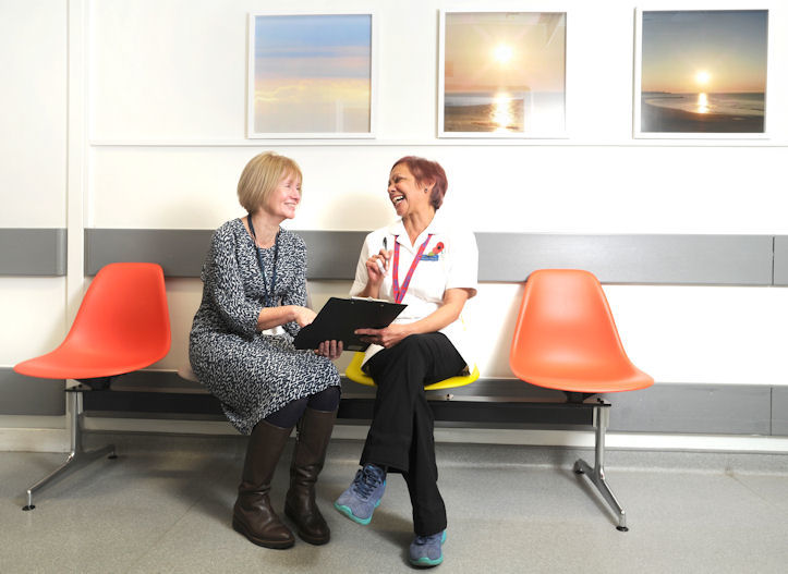Two smiling women with lanyards sit and chat on plastic chairs in a corridor with three sunset pictures on the wall behind