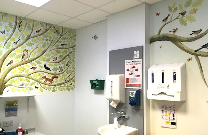 A hospital clinic room with wash basin and large naive image of oak tree and forest creatures on the wall
