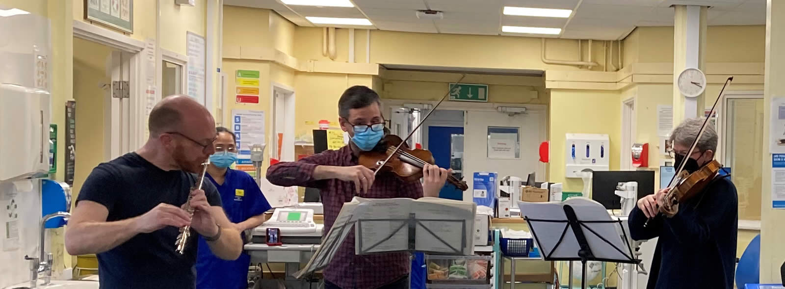 Three musicians play flute and violins in a hospital ward setting