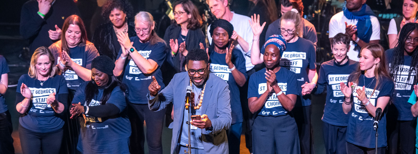 Around 20 applauding performers on stage in matching T-shirts; man in casual suit at front, hand raised, looking at mobile phone