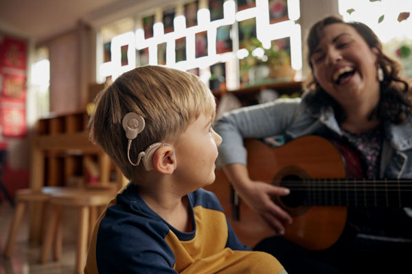 Small boy with cochlear implant device on his head sits in nursery classroom smiling at laughing young woman playing guitar