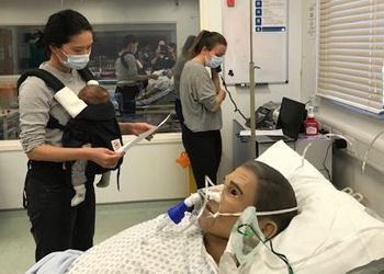 A dummy in a patient bed on a ward attended by two young people, one with an infant in a carrier on her chest.