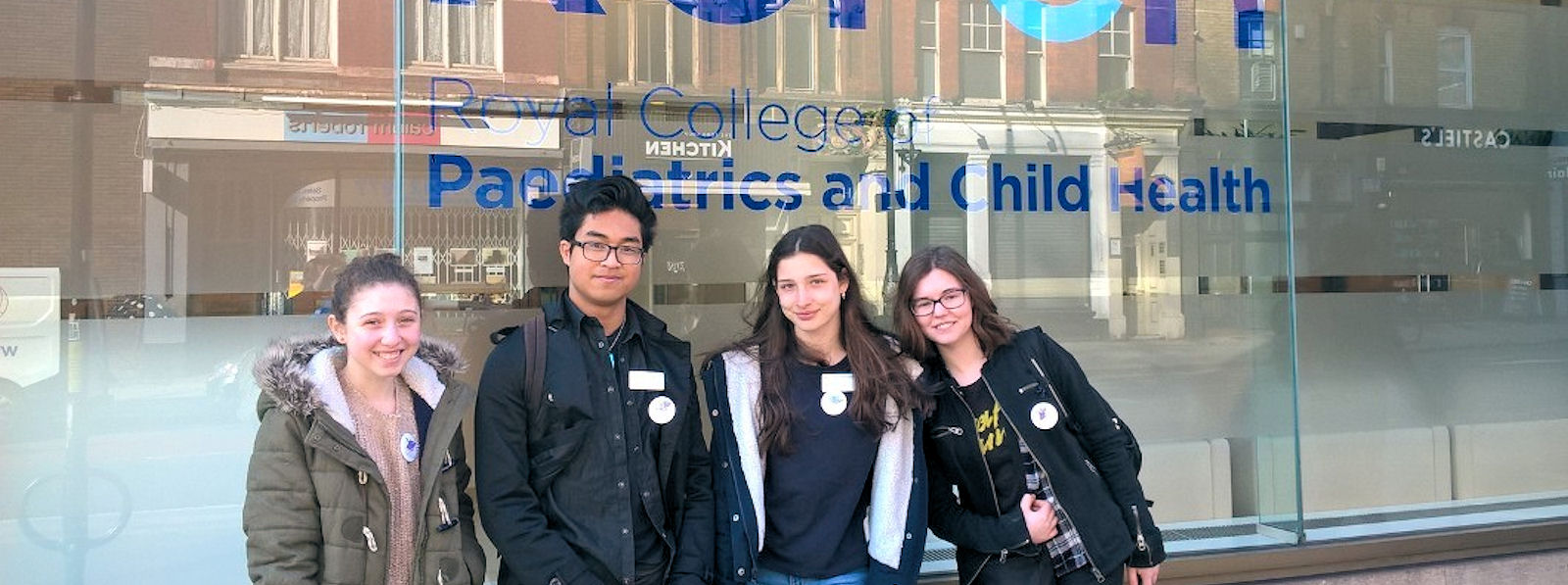 Four teenagers in warm jackets with name tags, in a row in front of the window of the Royal College of Paediatrics and Child Health