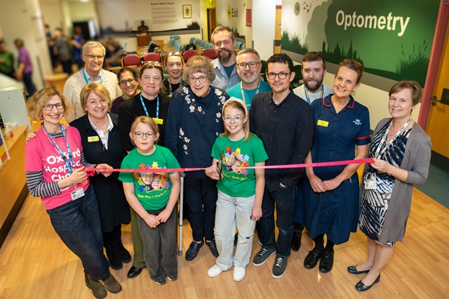 Group of people behind a ribbon. There is a new 'Optometry' wall design in the background. One person wears a pink 'Oxford Hospitals Charity' t-shirt.