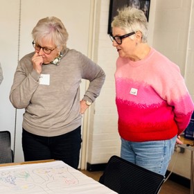 Two older women with name stickers examine the corner of a sheet on a table where there is a felt-tip drawing of lungs.
