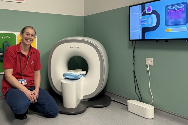 A member of hospital staff kneels beside a child‑friendly miniature MRI scanner known as a “kitten scanner” in a hospital play room. The small scanner is positioned on the floor, with a screen on the wall showing a cartoon animation designed to explain an MRI scan to children.