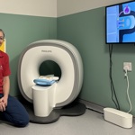 A member of hospital staff kneels beside a child‑friendly miniature MRI scanner known as a “kitten scanner” in a hospital play room. The small scanner is positioned on the floor, with a screen on the wall showing a cartoon animation designed to explain an MRI scan to children.