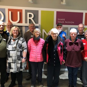 10 smiling middle-aged people stand in front of a wall with a bright design and 'Share your passion' written on it