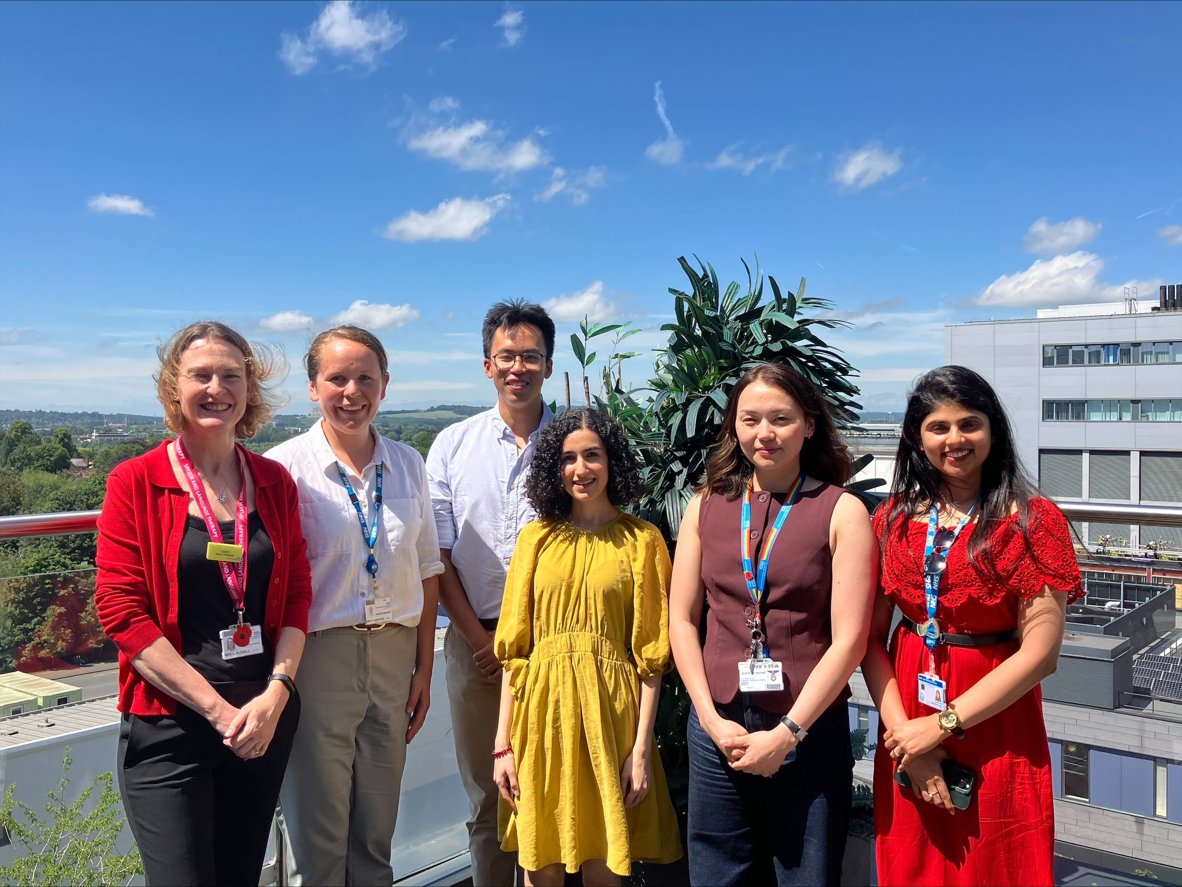 six people - five women and one man - facing the camera, with buildings and blue sky behind them