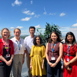 six people - five women and one man - facing the camera, with buildings and blue sky behind them