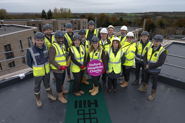 Group of 15 people on a roof and wearing PPE (helmets and high visisiubility jackets). One person holds a banner with the text 'Oxford Hospitals Charity'). Completed buildings are in the background.