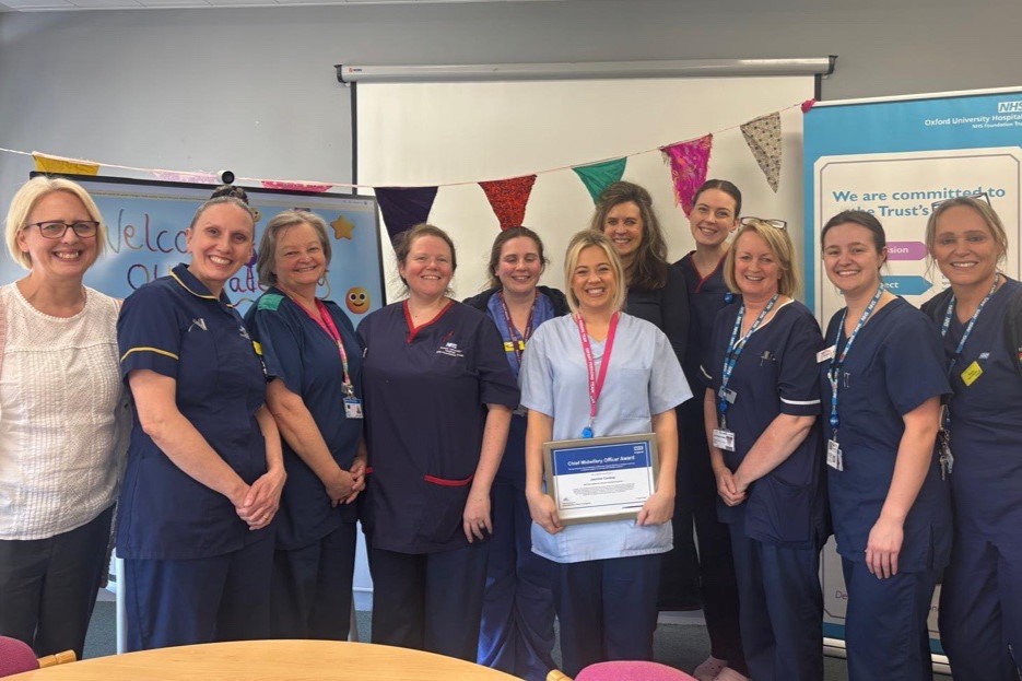 Eleven NHS staff stand together in a meeting room, wearing clinical uniforms. One person (Jasmin) holds a certificate in the centre of the group. Oxford University Hospitals branding and bunting are displayed behind them.