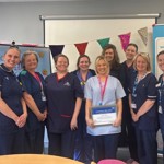 Eleven NHS staff stand together in a meeting room, wearing clinical uniforms. One person (Jasmin) holds a certificate in the centre of the group. Oxford University Hospitals branding and bunting are displayed behind them.