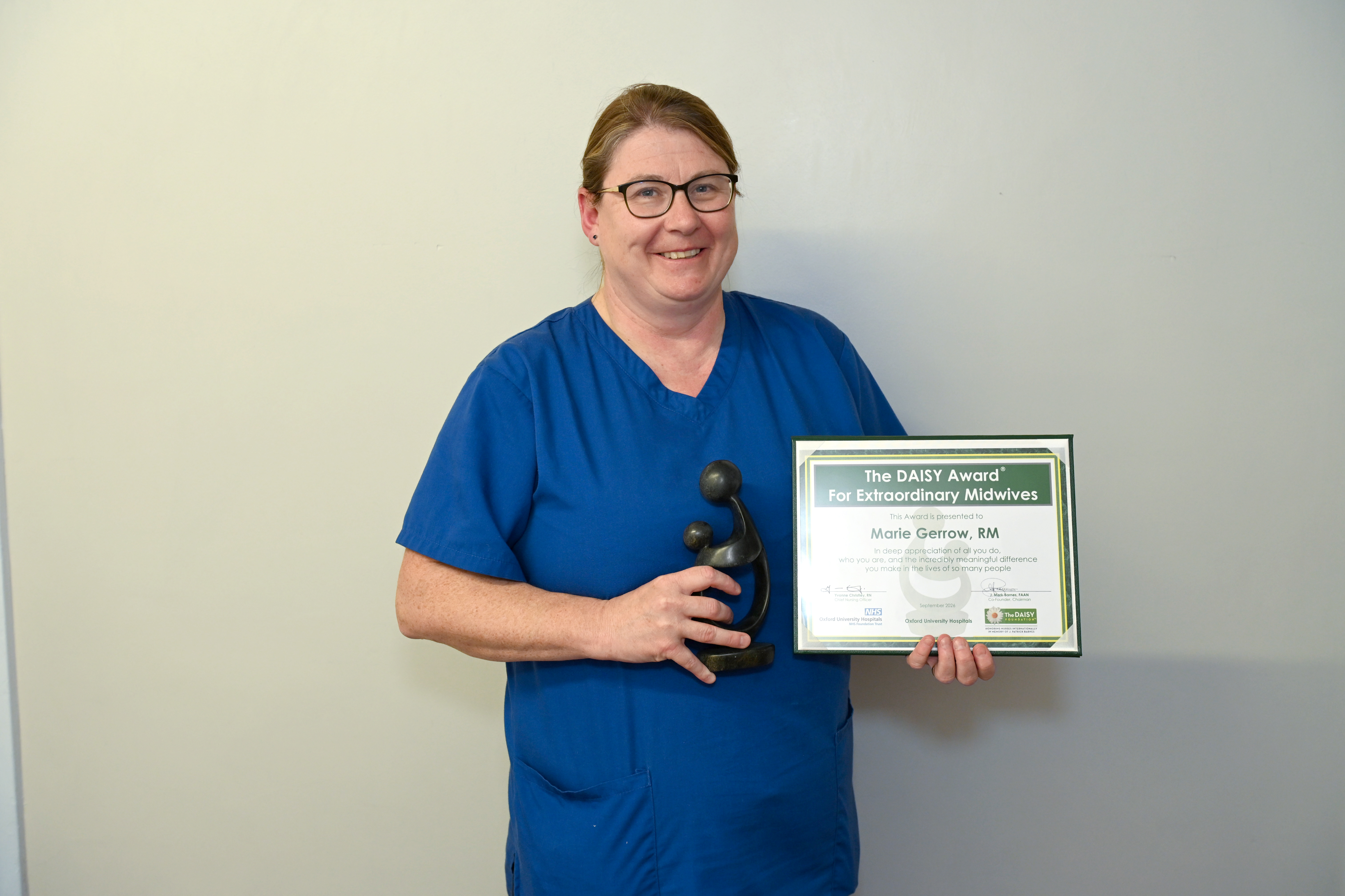 Midwife in blue scrubs holding a DAISY Award certificate and trophy