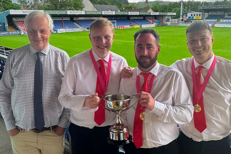 Four men standing at a football stadium, three are wearing red ties and medals. The middle two have a hand each on a trophy