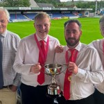 Four men standing at a football stadium, three are wearing red ties and medals. The middle two have a hand each on a trophy