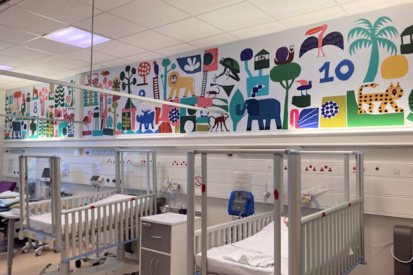 Row of cots in a ward beneath a long wall painted with naive colourful animals, birds and trees.