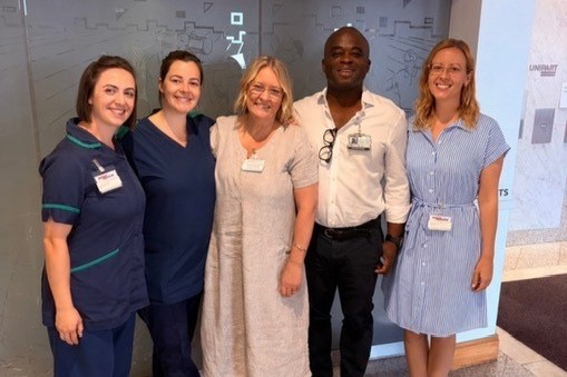 Five people standing together indoors in a corridor, posing for a group photo. Two on the left are wearing clinical uniforms.