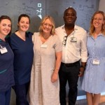Five people standing together indoors in a corridor, posing for a group photo. Two on the left are wearing clinical uniforms.