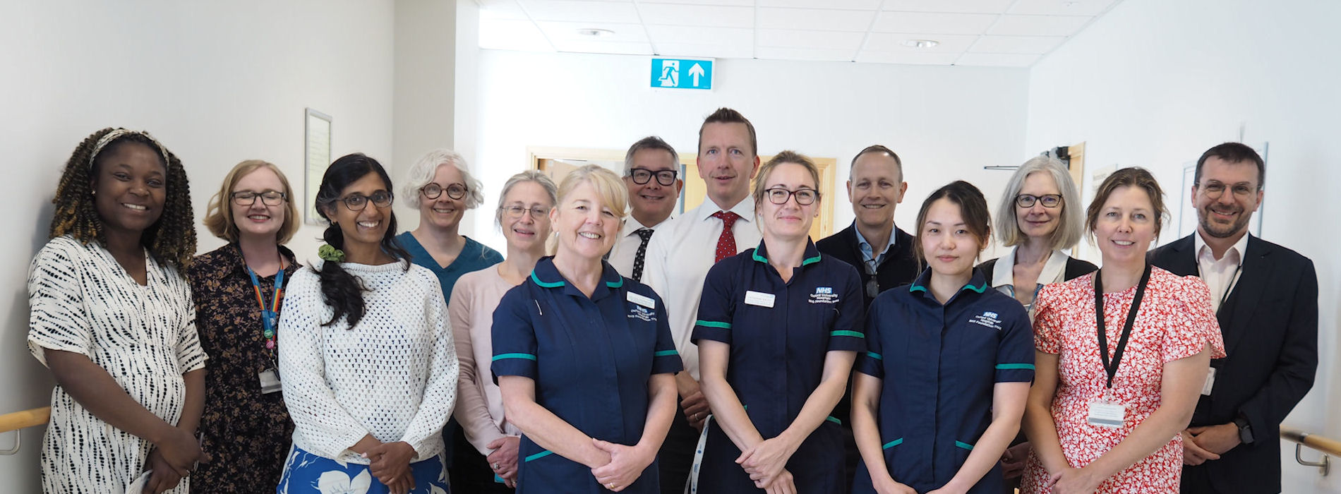 Ten women and four men in smart dress or healthcare uniforms stand smiling in a bright hospital room.