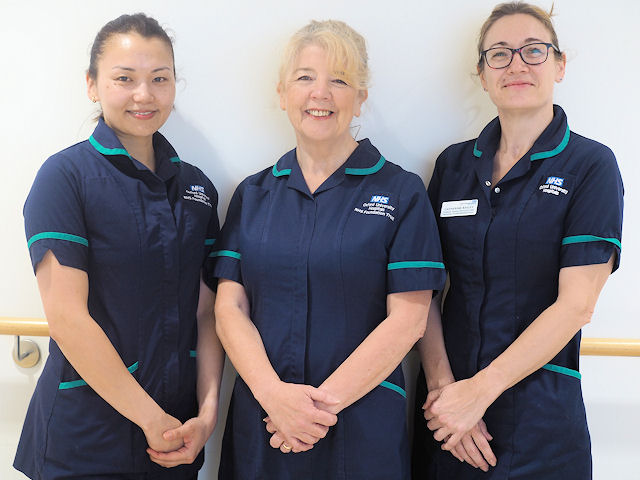 Three smiling women in nurses' uniforms, hands clasped in front.