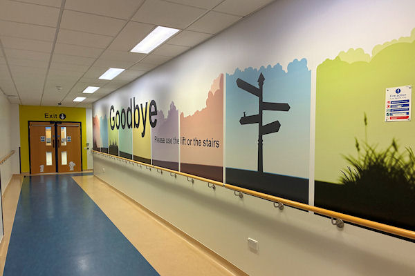 Hospital corridor with double door at end; colourful wall panels with word 'Goodbye' and old-fashioned signpost depicted.