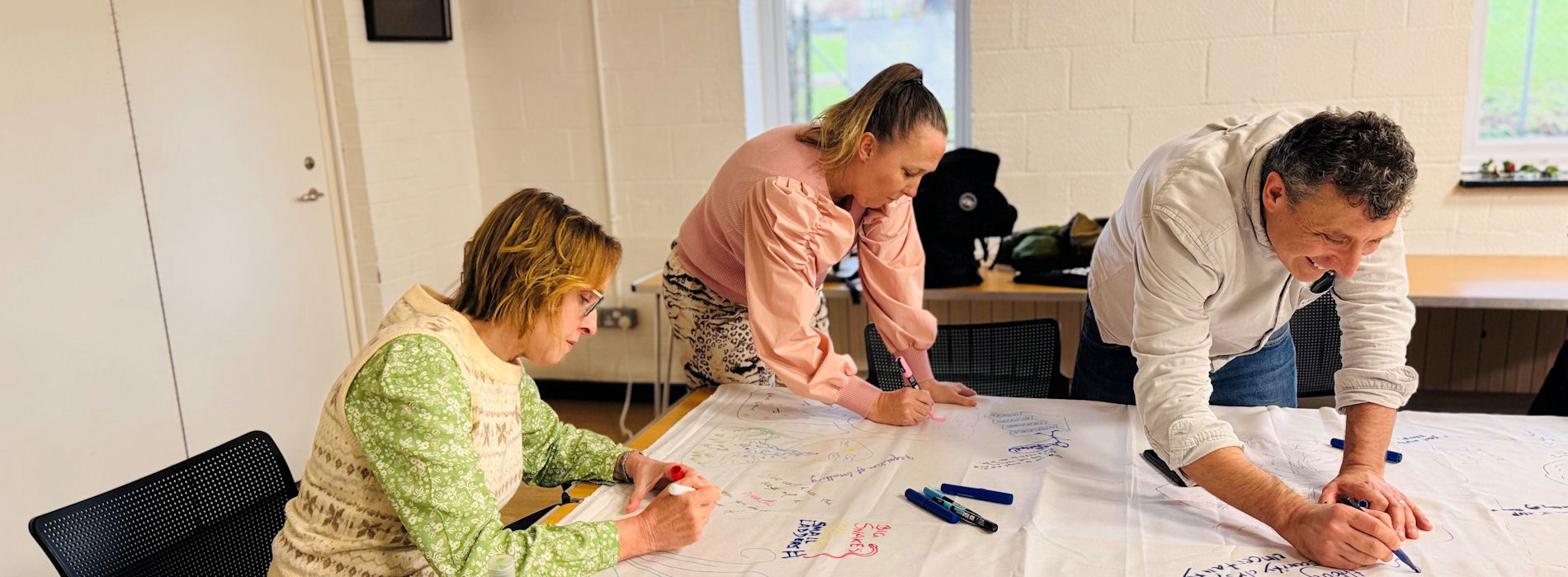 A man and two women in long-sleeved shirts lean over and write on a paper-covered table.