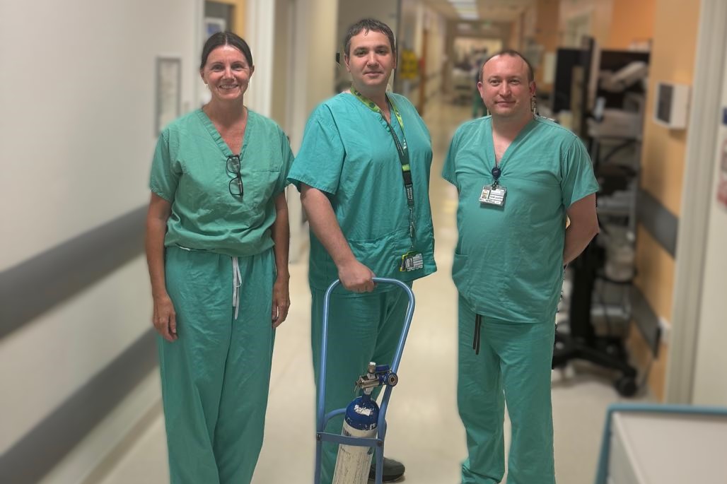 Three healthcare staff in green scrubs stand in a hospital corridor beside an oxygen cylinder