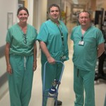 Three healthcare staff in green scrubs stand in a hospital corridor beside an oxygen cylinder