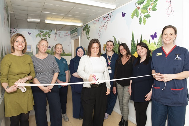 A group of staff members stand together in a hospital corridor decorated with nature-themed wall art. The person in the centre cuts a ceremonial ribbon.