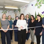 A group of staff members stand together in a hospital corridor decorated with nature-themed wall art. The person in the centre cuts a ceremonial ribbon.