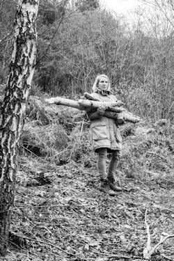Woman in anorak carries pile of logs in birch wood clearing