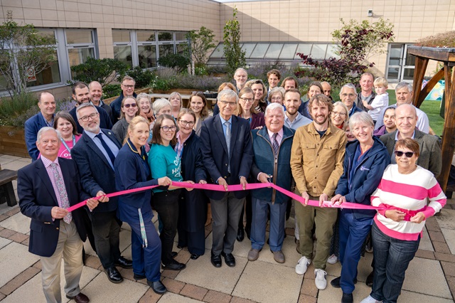 Big group of people, including uniformed members of staff and people wearing 'Oxford Hospitals Charity' t-shirts. Many are smiling. People at the front are holding a pink ribbon and a person is about to cut it.