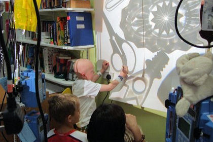 Three children, one bald, interact with shadow images on a large wall screen, cluttered bookshelf in background.