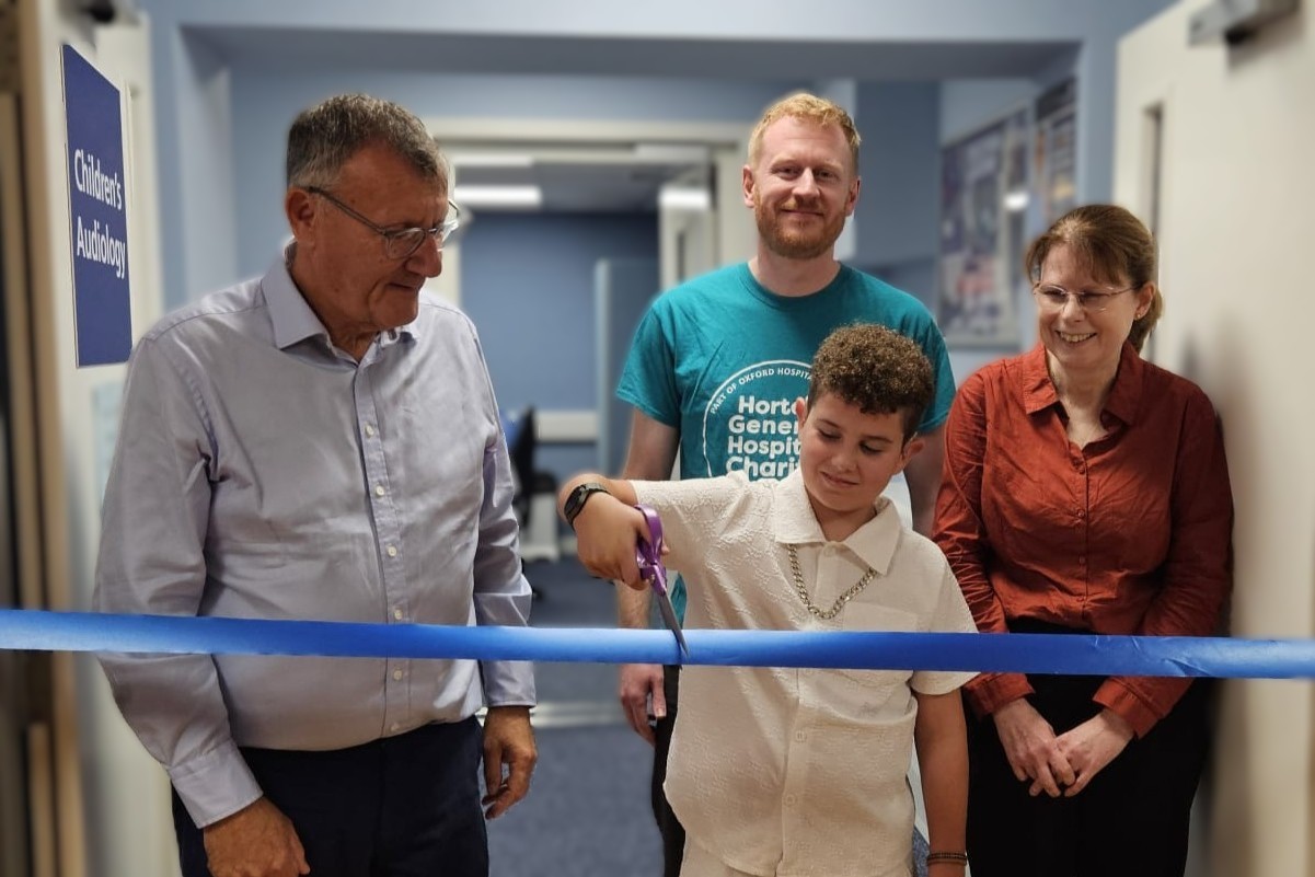 A child cuts a blue ribbon at a hospital audiology department, with three people standing alongside.