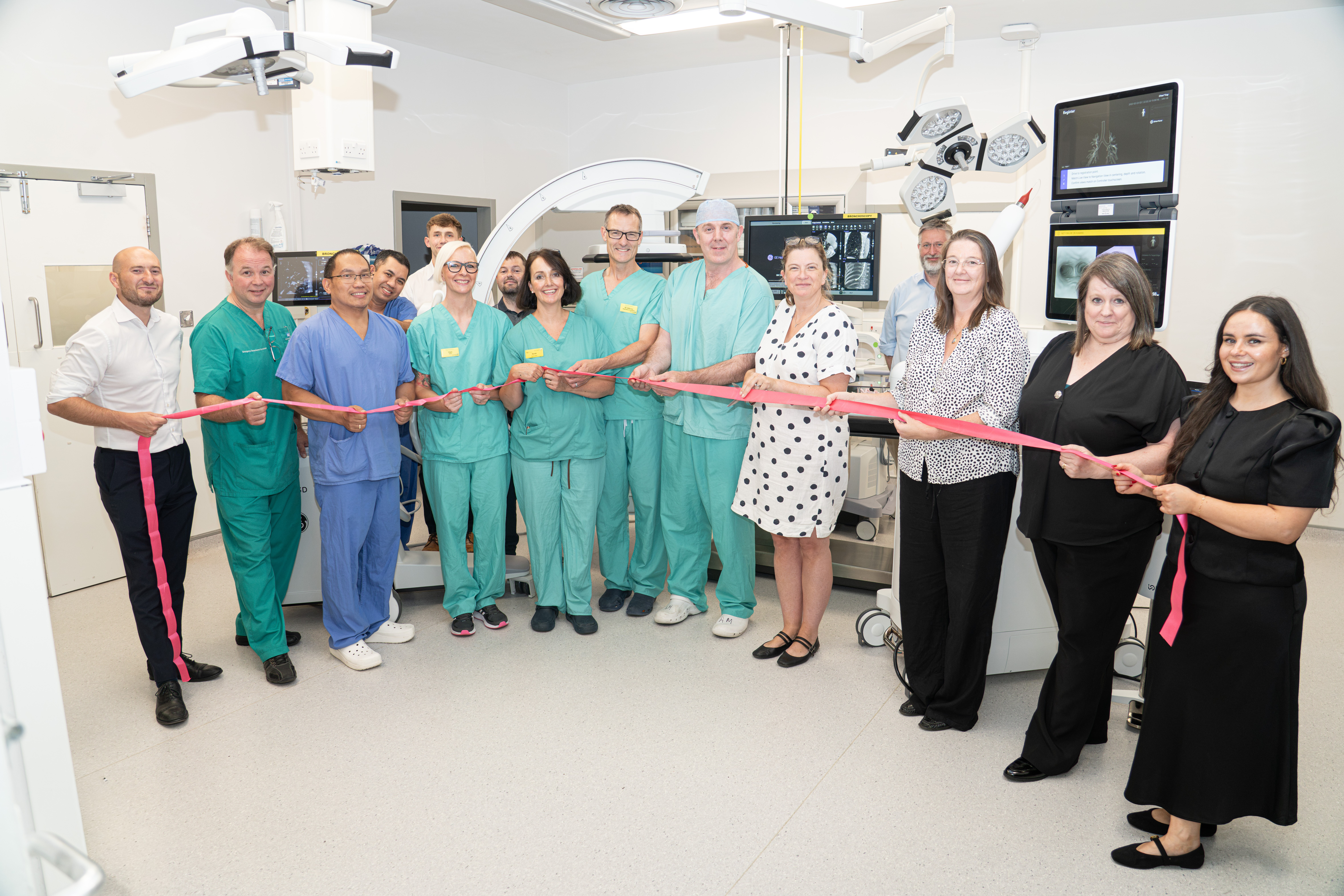 Group of 15 people, including some in clinical uniform, pose for a ribbon cutting in a clinical hospital room. The equipment is behind most of the people.