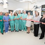 Group of 15 people, including some in clinical uniform, pose for a ribbon cutting in a clinical hospital room. The equipment is behind most of the people.