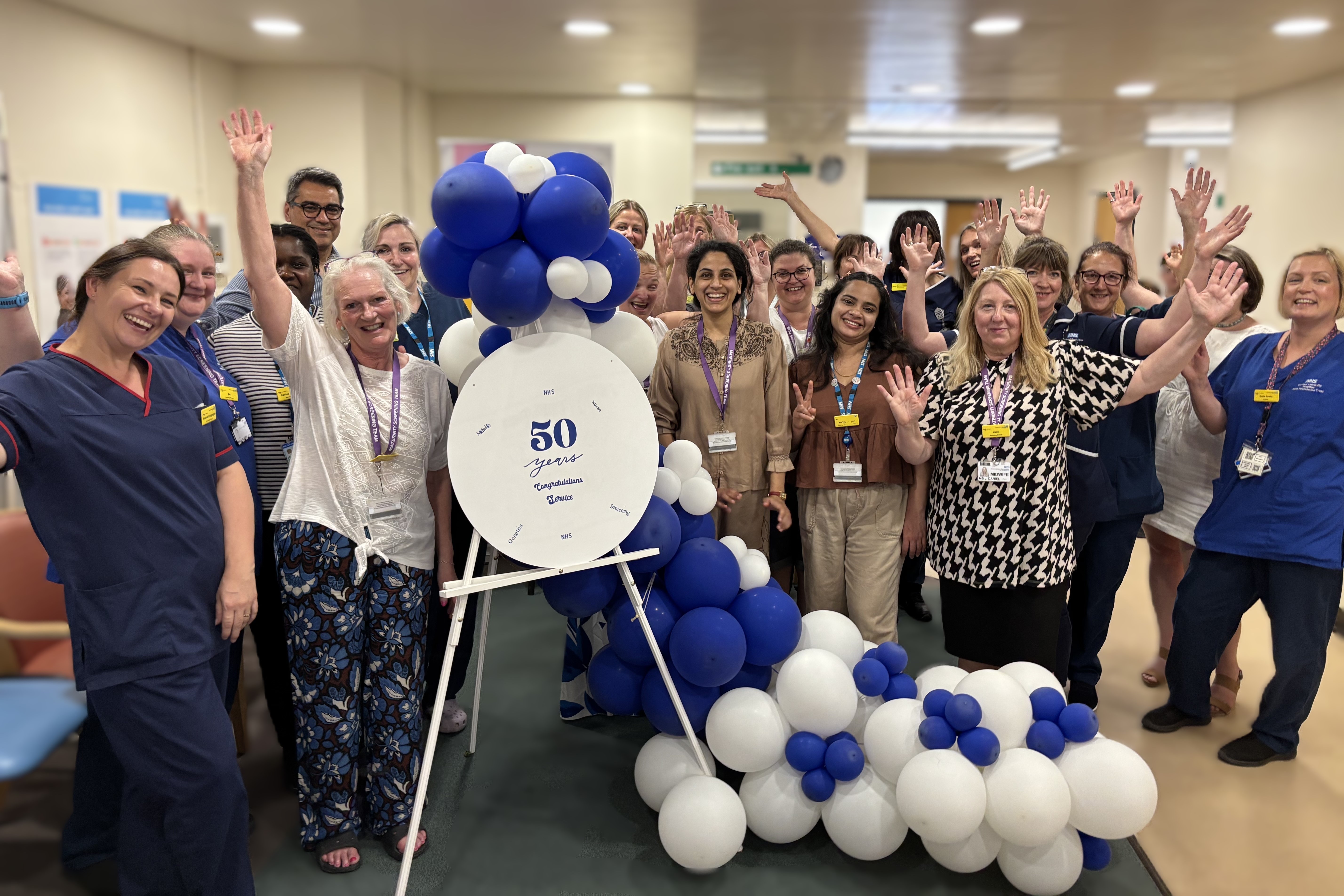 Group of 21 maternity staff gathered around a “50 years of service” sign and blue-and-white balloons, raising their hands in celebration.