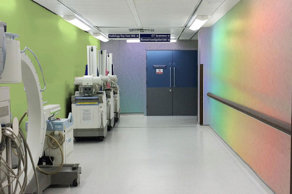 Hospital corridor with equipment to left and double doors at end, rainbow coloured gradient on right-hand wall.
