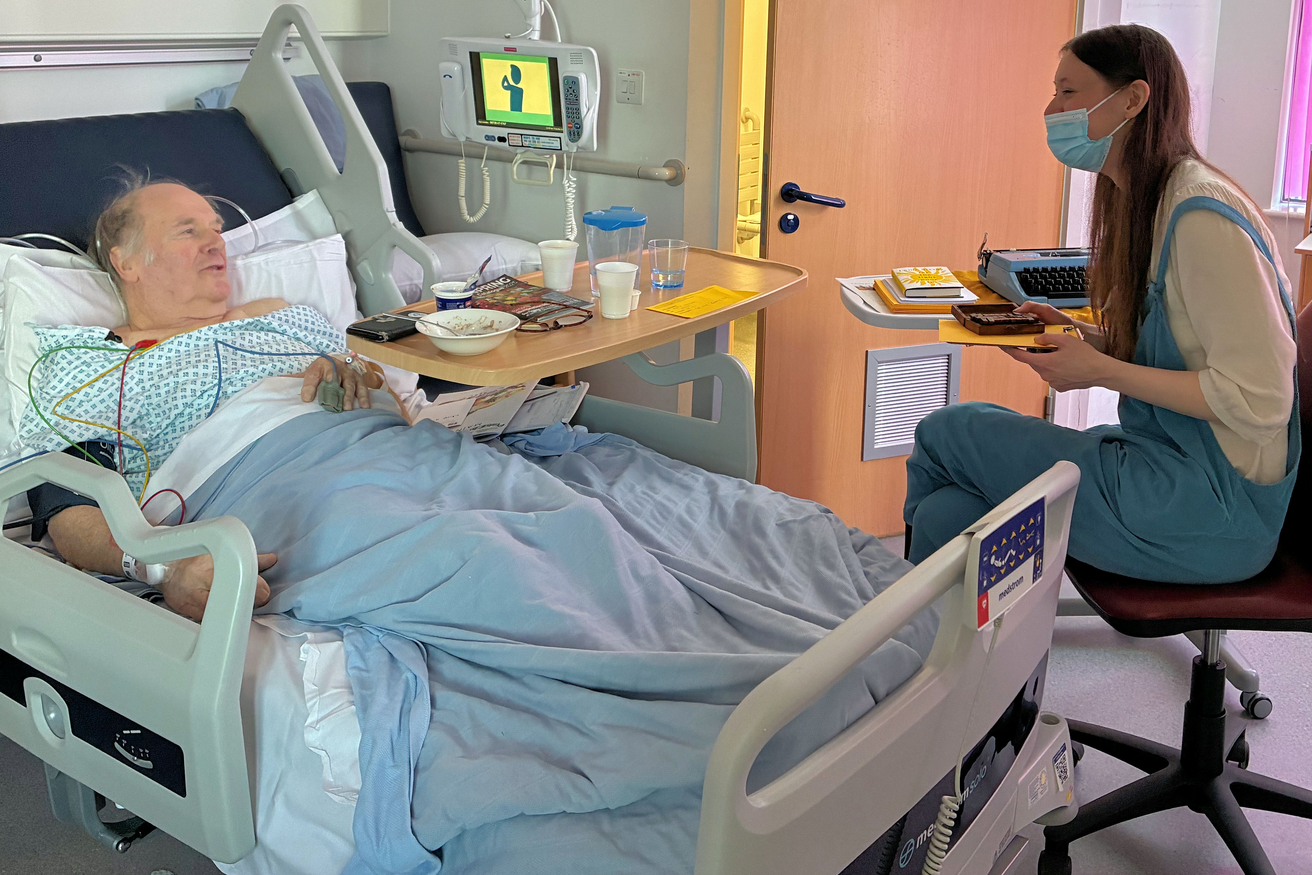 Elderly man in hospital bed speaks to young woman in dungarees and face mask.