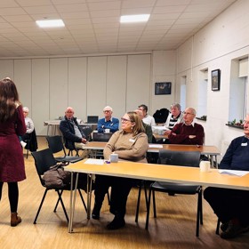 Around 10 people seated at long tables listening to a young female speaker with her back to the camera.