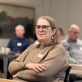 Woman with name sticker 'Lyn', arms folded, seated at a table and smiling, three indistinct figures behind.