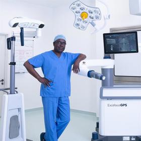 In a clean surgical setting a relaxed, confident man in scrubs leans on a large clinical machine labelled 'ExcelsisusGPS'. It has a display screen and a theatre light above it.
