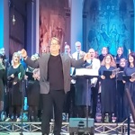 A conductor leading a large choir performing on stage inside a church
